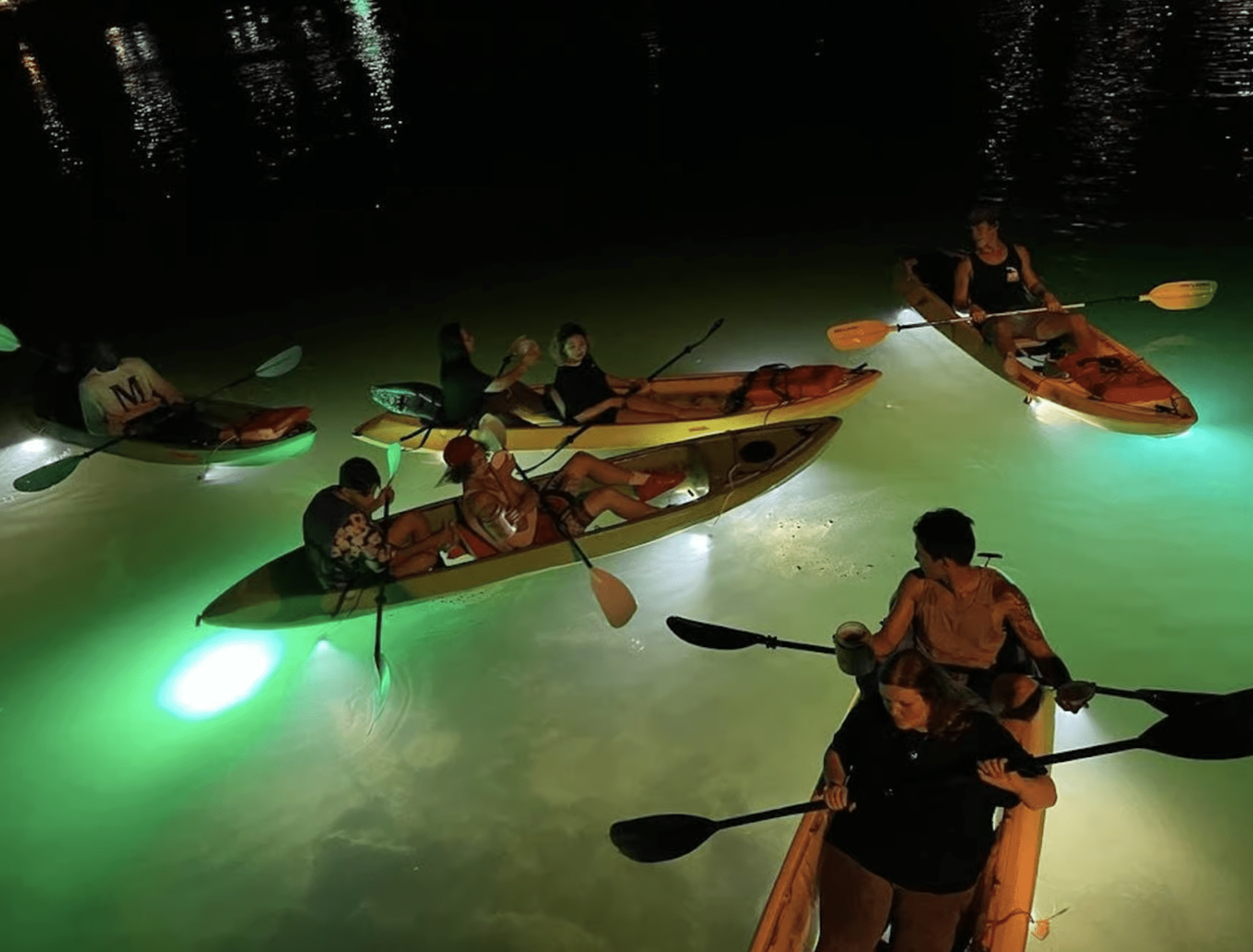 Group kayaking at night in clear kayaks illuminated by glowing underwater LED lights.