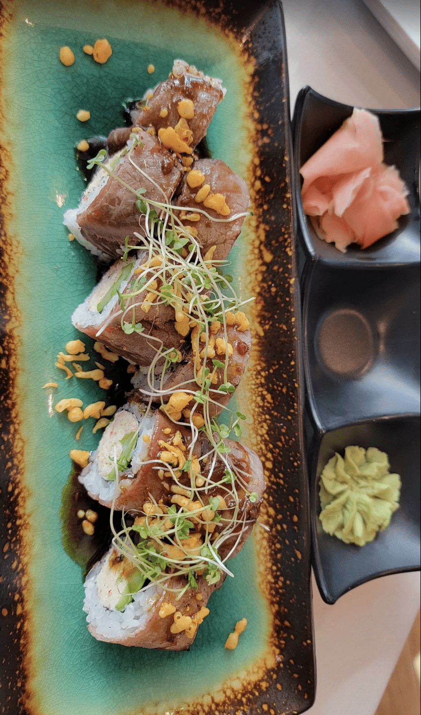 Sushi rolls topped with seared fish, microgreens, and crunchy bits, served with pickled ginger.